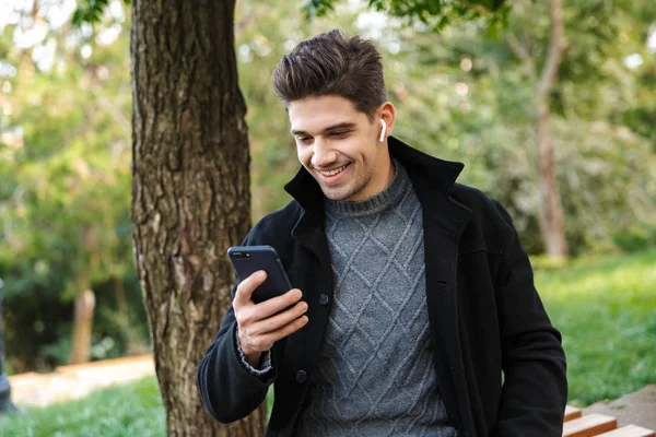 Glimlachend knappe jonge vrolijke man in Casual kleding wandelen buitenshuis in Green Park met behulp van mobiele telefoon luisteren muziek met oortelefoons. — Stockfoto