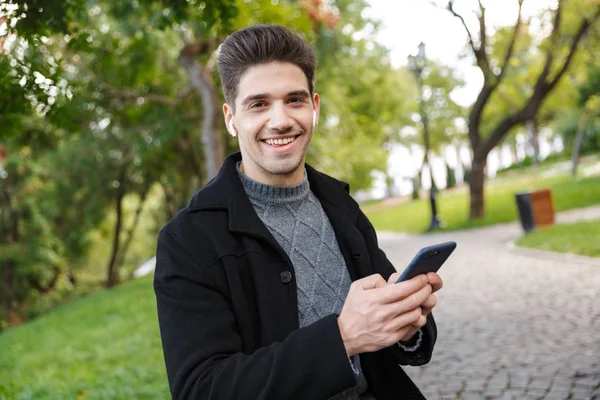 Mosolygós csinos fiatal vidám férfi alkalmi ruházati cikkek séta a szabadban, zöld Park segítségével mobiltelefon hallgató zene fülhallgató. — Stock Fotó