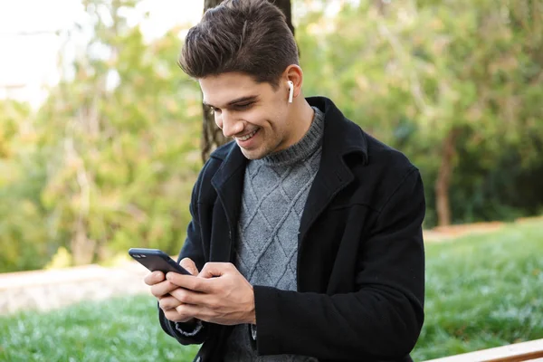 Glimlachend knappe jonge vrolijke man in Casual kleding wandelen buitenshuis in Green Park met behulp van mobiele telefoon luisteren muziek met oortelefoons. — Stockfoto