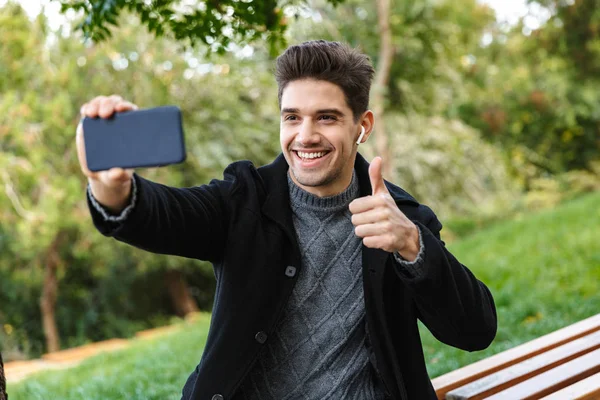 Joven optimista en ropa casual caminando al aire libre en el parque verde utilizando el teléfono móvil tomar una selfie con los pulgares hacia arriba gesto . — Foto de Stock