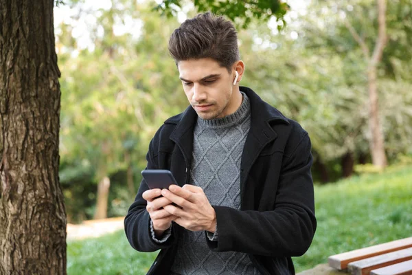 Concentrated handsome young man in casual clothing walking outdoors in green park using mobile phone listening music with earphones. — Stock Photo, Image