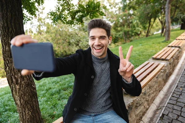 Allegro ragazzo contento in abbigliamento casual camminare all'aperto nel parco verde utilizzando il telefono cellulare farsi un selfie con gesto di pace . — Foto Stock