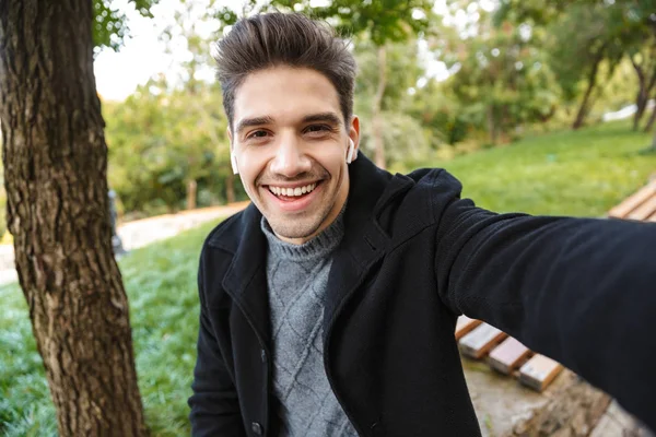 Lächelnder junger Mann in legerer Kleidung, der draußen im grünen Park spaziert, macht ein Selfie mit der Kamera. — Stockfoto