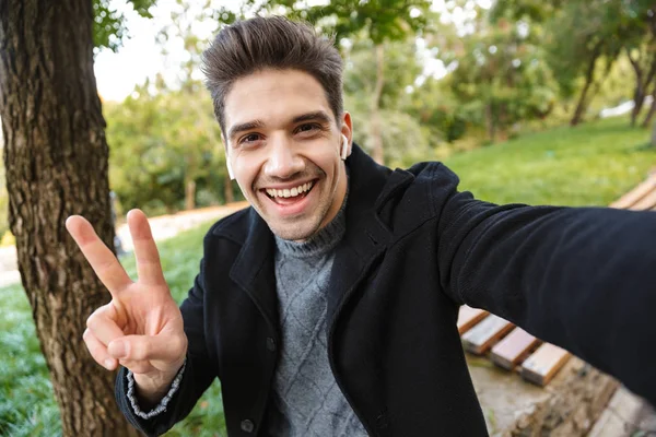 Fröhlicher junger Mann in lässiger Kleidung, der draußen im grünen Park spaziert, macht ein Selfie mit der Kamera und hört Musik mit Kopfhörern. — Stockfoto