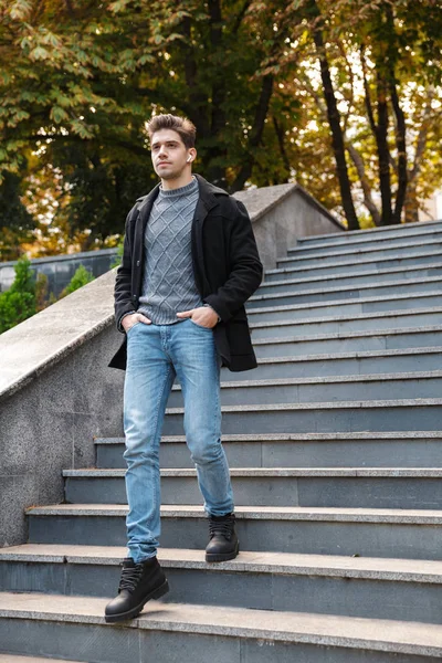 Young man in casual clothing walking outdoors in green park by steps listening music with earphones. — Stock Photo, Image