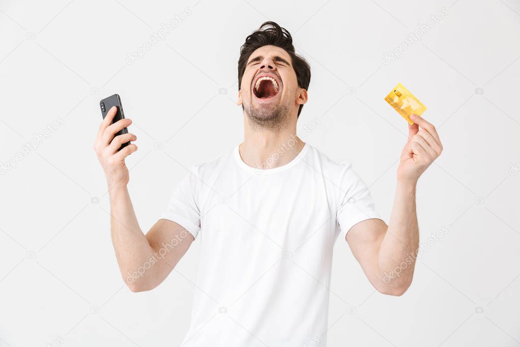 Excited happy young man posing isolated over white wall background using mobile phone holding credit card.