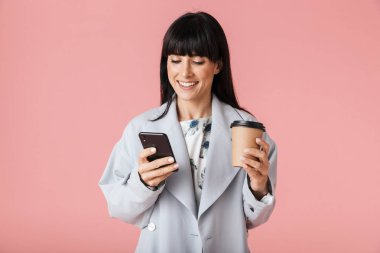 Kahve tutan cep telefonu kullanarak açık pembe arka plan duvarında izole poz kadın.