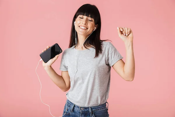 Bella giovane donna felice posa isolata su sfondo rosa parete ascoltare musica con auricolari . — Foto Stock