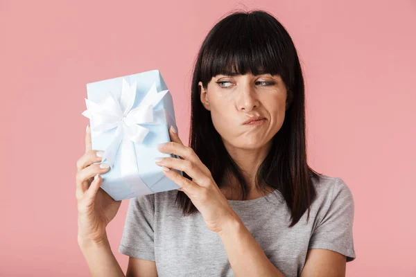 Beautiful amazing confused thinking woman posing isolated over light pink background wall holding present gift box. — Stock Photo, Image