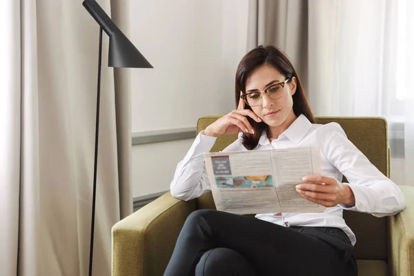 Concentrated beautiful young business woman in formal wear clothes indoors at home reading newspaper.