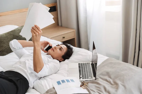 Hermosa joven mujer de negocios en ropa de vestir formal en el interior de casa se encuentra en la cama hablando por teléfono sosteniendo documentos en papel . —  Fotos de Stock