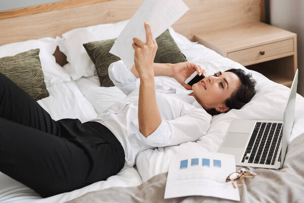 Beautiful young business woman in formal wear clothes indoors at home lies on bed talking by phone holding paper documents.