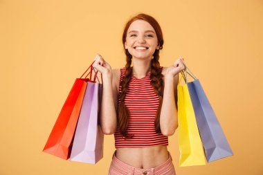 Mutlu genç güzel kızıl saçlı kadın alışveriş çantaları tutarak sarı arka plan üzerinde izole poz.