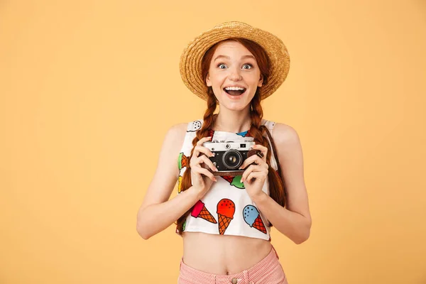 Ευτυχισμένος νέος όμορφη κοκκινομάλλα γυναίκα φωτογράφος τουρίστας ντυμένος στο παγωτό τυπωμένο μπλουζάκι που παρουσιάζουν απομονωμένη πάνω από κίτρινο φόντο κρατώντας φωτογραφική μηχανή. — Φωτογραφία Αρχείου