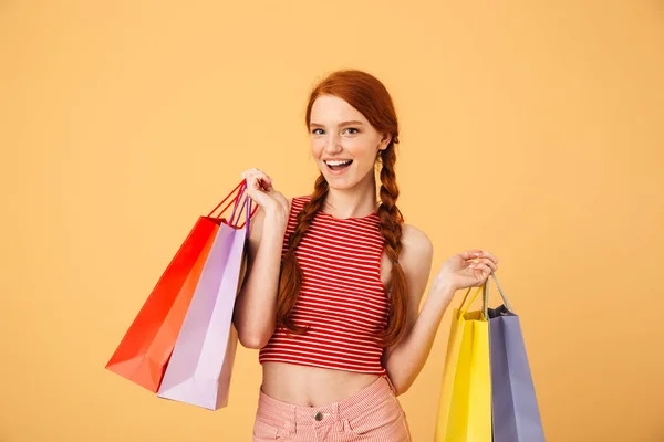 Agradable joven bonita pelirroja posando aislada sobre fondo amarillo sosteniendo bolsas de compras . — Foto de Stock