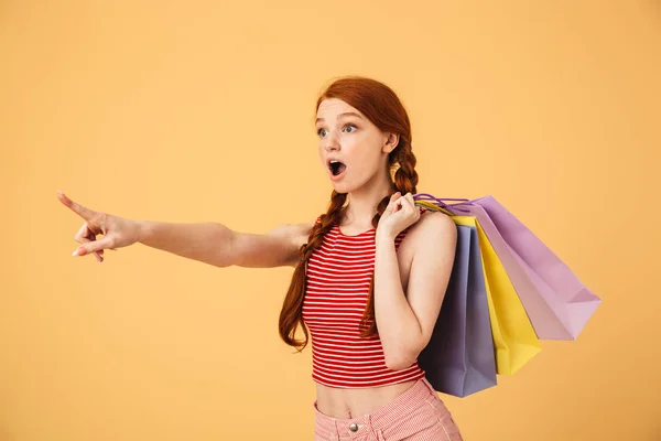 Sorprendido joven hermosa pelirroja posando aislado sobre fondo amarillo sosteniendo bolsas de compras . — Foto de Stock
