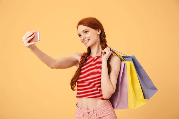 Uśmiechnięta młoda Ładna Ruda kobieta pozowanie na białym tle żółte tło trzyma torby na zakupy zrobić selfie przez telefon komórkowy. — Zdjęcie stockowe