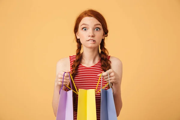 Νευρική ενθουσιασμένη νεαρή όμορφη κοκκινομάλλα γυναίκα που θέτει απομονωμένοι πάνω από κίτρινο φόντο κρατώντας σακούλες αγορών αναζητούν φωτογραφική μηχανή. — Φωτογραφία Αρχείου