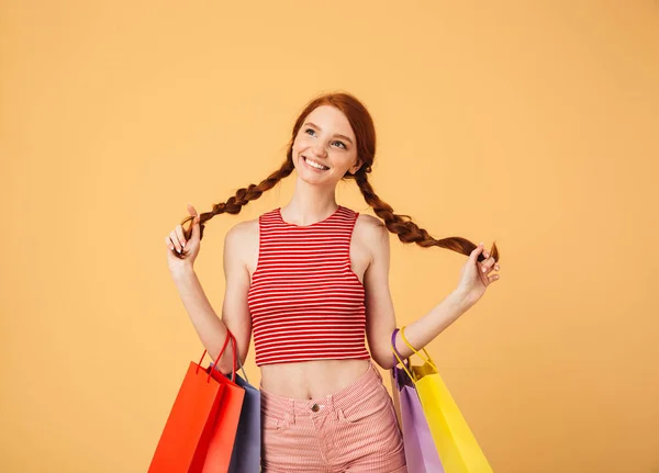 Linda sonriente joven bonita pelirroja posando aislada sobre fondo amarillo sosteniendo bolsas de compras . — Foto de Stock