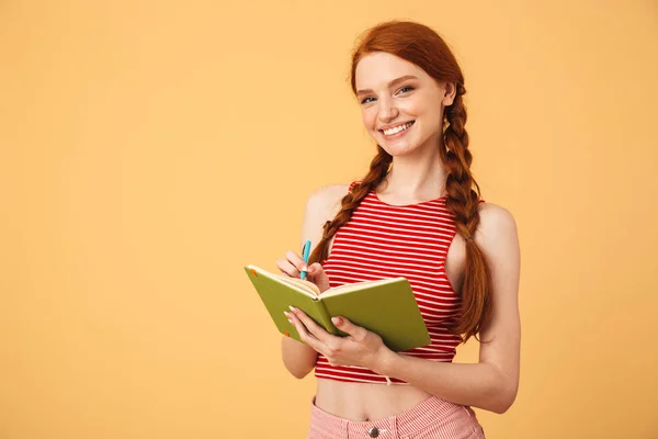 Cheerful  young beautiful redhead woman posing isolated over yellow background holding book reading. — Stock Photo, Image