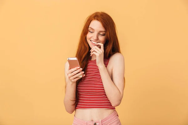 Усміхнений задоволений милий молодий красива руда жінка позує ізольовано на жовтому тлі за допомогою мобільного телефону балачки . — стокове фото