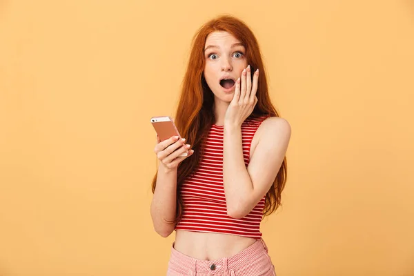 携帯電話のチャットを使用して黄色の背景の上に孤立したポーズの驚いた感情的なかわいいかわいい赤毛の女性. — ストック写真