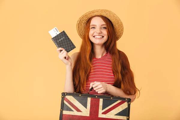 Cute young pretty redhead woman posing isolated over yellow background holding suitcase and passport with tickets — Stock Photo, Image