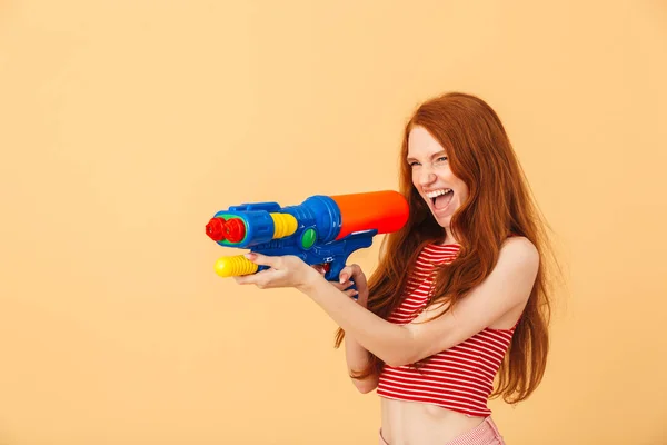 Возбужденная эмоциональная молодая красивая рыжая женщина позирует изолированно на желтом фоне держа игрушку водяного пистолета . — стоковое фото