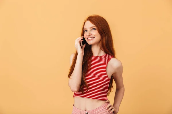 Задоволена молода красива руда жінка позує ізольовано на жовтому фоні, розмовляючи мобільним телефоном . — стокове фото