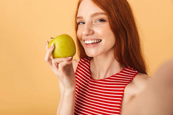 Szczęśliwy młody piękny rudowłosy kobieta stwarzające izolowane na żółtym tle wziąć selfie przez aparat gospodarstwa jabłko. — Zdjęcie stockowe