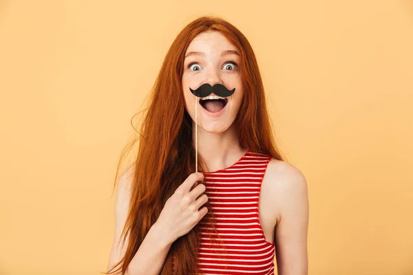 Choqué émotionnel jeune belle rousse femme posant isolé sur fond jaune tenant fausse moustache . — Photo