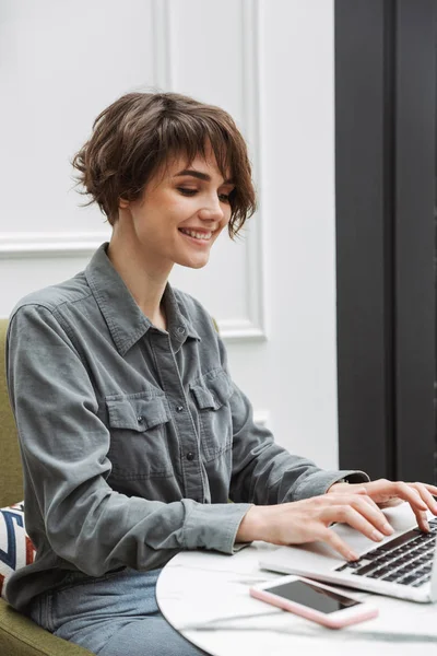 Jovem feliz estudante de negócios bonita sentada no café dentro de casa usando computador portátil . — Fotografia de Stock