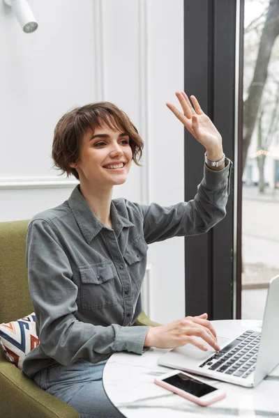 Estudante de negócios mulher sentada no café dentro de casa usando computador portátil coworking acenando para os amigos . — Fotografia de Stock