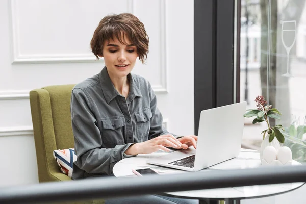 Fornøyd ung, pen student kvinne som sitter på kafeen innendørs med laptop-coworking og ser på mobiltelefon . – stockfoto