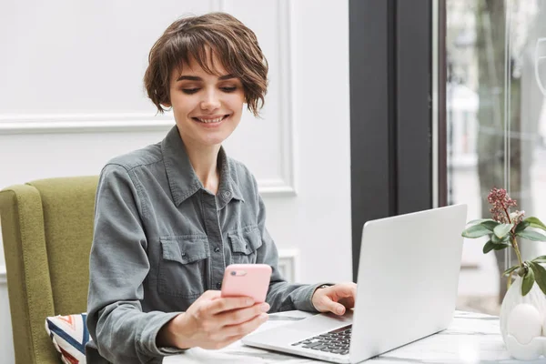 携帯電話でおしゃべりするノートパソコンを使って屋内のカフェに座っている若いかわいいビジネス学生の女性. — ストック写真