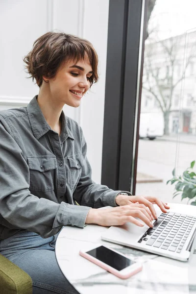 Jovem aluna de negócios bonita alegre sentada no café dentro de casa usando computador portátil coworking . — Fotografia de Stock