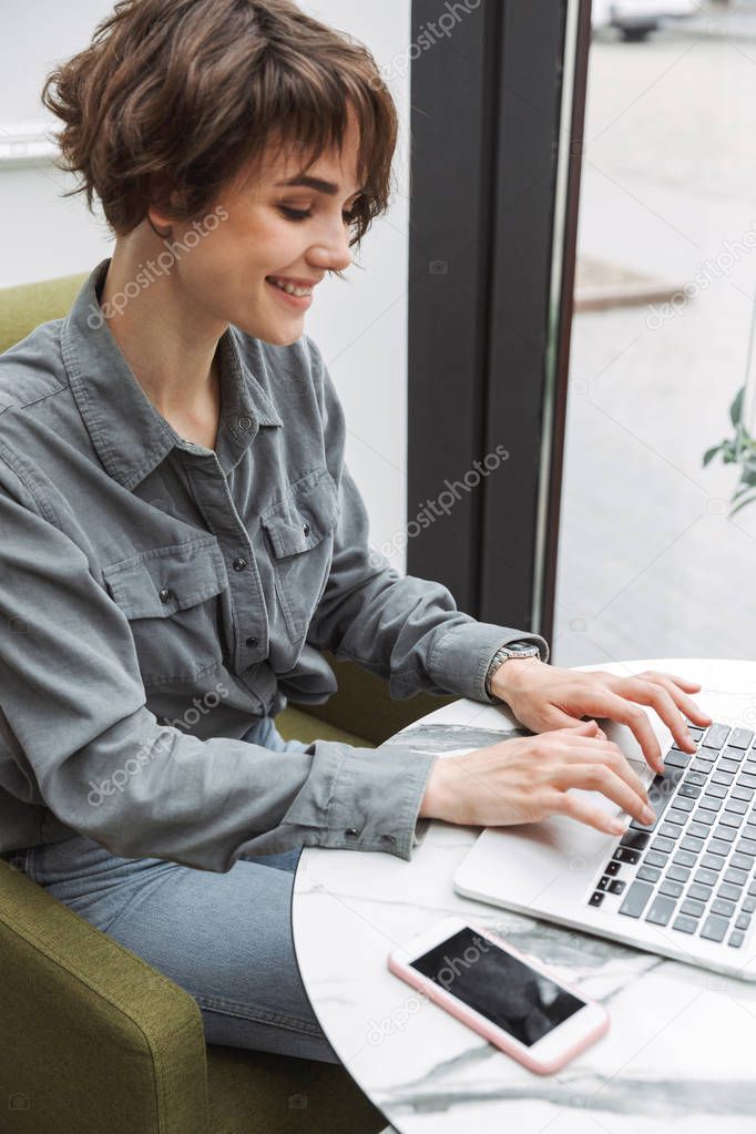 Happy cheerful young pretty business student woman sitting in cafe indoors using laptop computer coworking.