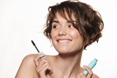 Sağlıklı parlak cilt ile Neşeli genç güzel kadın beyaz duvar arka plan üzerinde izole poz kirpik maskara makyaj uygulamak tutarak.