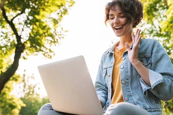 Bild einer zufriedenen kaukasischen Frau mit silbernem Laptop, während sie auf einer Bank im grünen Park sitzt — Stockfoto