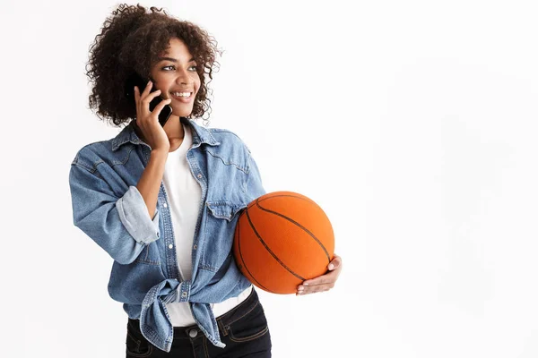 Mladá vzrušená sportovní žena Africká holdingová basketbalová, která se izoluje na pozadí bílé zdi mluvící mobilním telefonem. — Stock fotografie