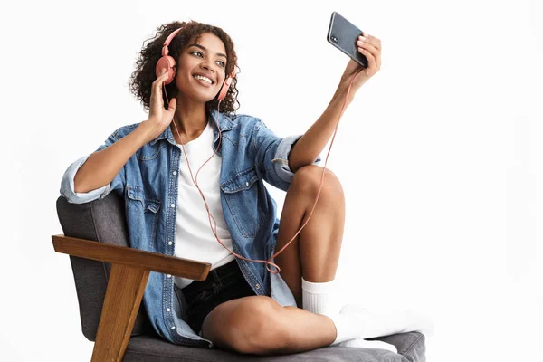 Upphetsad kvinna afrikansk lyssnar musik i hörlurar poserar isolerade över vit vägg bakgrund pratar med mobiltelefon ta en selfie. — Stockfoto