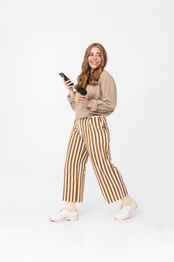 Neşeli gülümseyen sevimli genç genç kız kahve içme cep telefonu kullanarak beyaz duvar arka plan üzerinde izole yürüyüş.