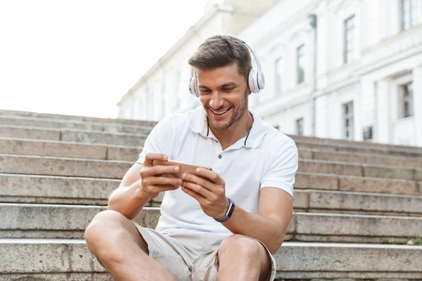 Retrato del joven alegre sonriendo y sosteniendo el teléfono inteligente mientras —  Fotos de Stock