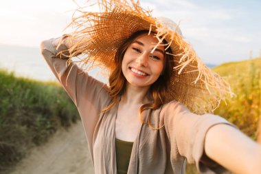Güzel neşeli mutlu gülümseyen genç güzel kadın plaj denizi yakınında doğa yeşil çim açık havada poz kamera ile selfie çekmek.