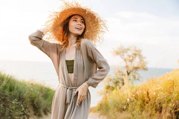Bastante alegre feliz sonriente joven mujer en sombrero posando al aire libre en la naturaleza hierba verde cerca de la playa mar . — Foto de Stock