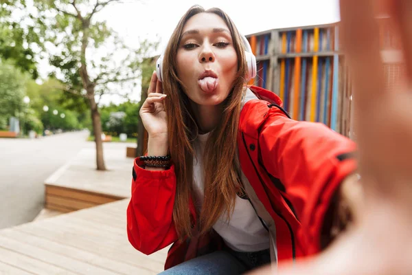Jeune jolie femme heureuse en imperméable posant à l'extérieur écoutant de la musique avec des écouteurs prendre selfie par caméra montrant la langue . — Photo