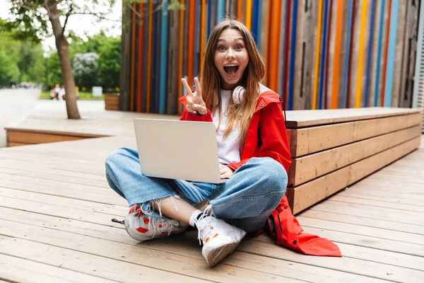 Beautiful young happy pretty woman in raincoat posing outdoors using laptop computer showing peace gesture. — Stock Photo, Image