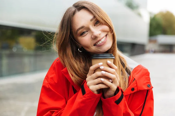 Mooie opgewonden emotionele jonge gelukkig mooie vrouw in regenjas poseren buitenshuis drinken koffie. — Stockfoto