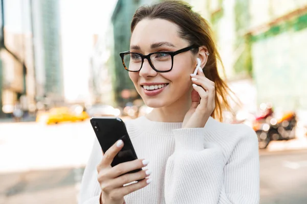 Foto close-up de mulher muito sorridente usando celular e fone de ouvido enquanto caminhava na rua da cidade grande — Fotografia de Stock