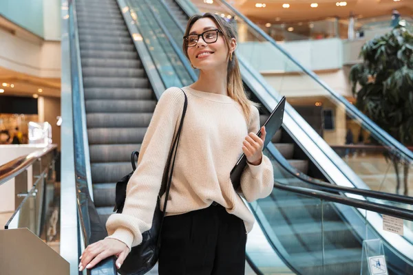 Foto de una linda mujer joven sonriendo y sosteniendo el portapapeles mientras baja por la escalera mecánica en el edificio interior — Foto de Stock